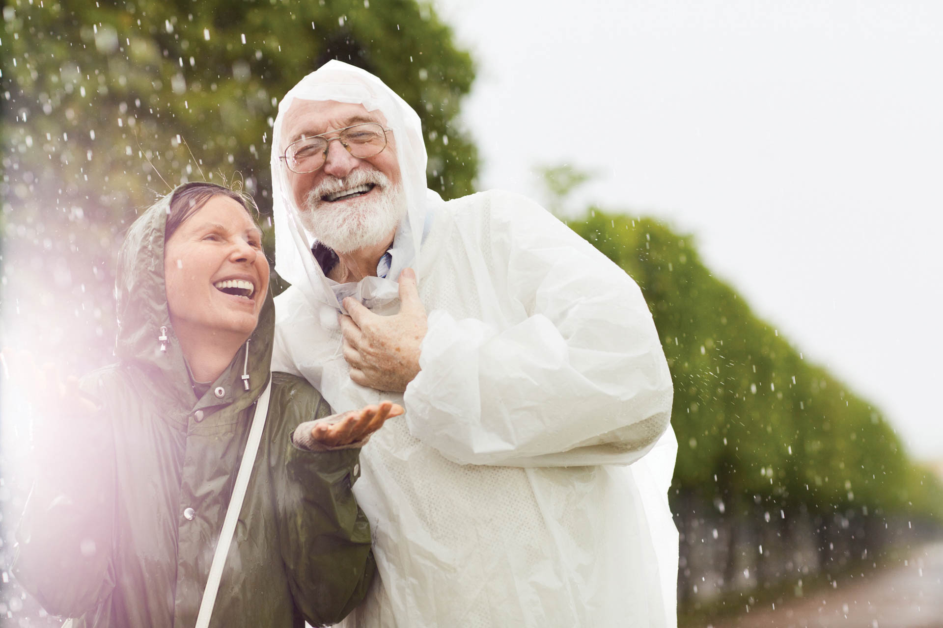 Cheerful aged woman catching raindrops on palms with her laughing husband near by.