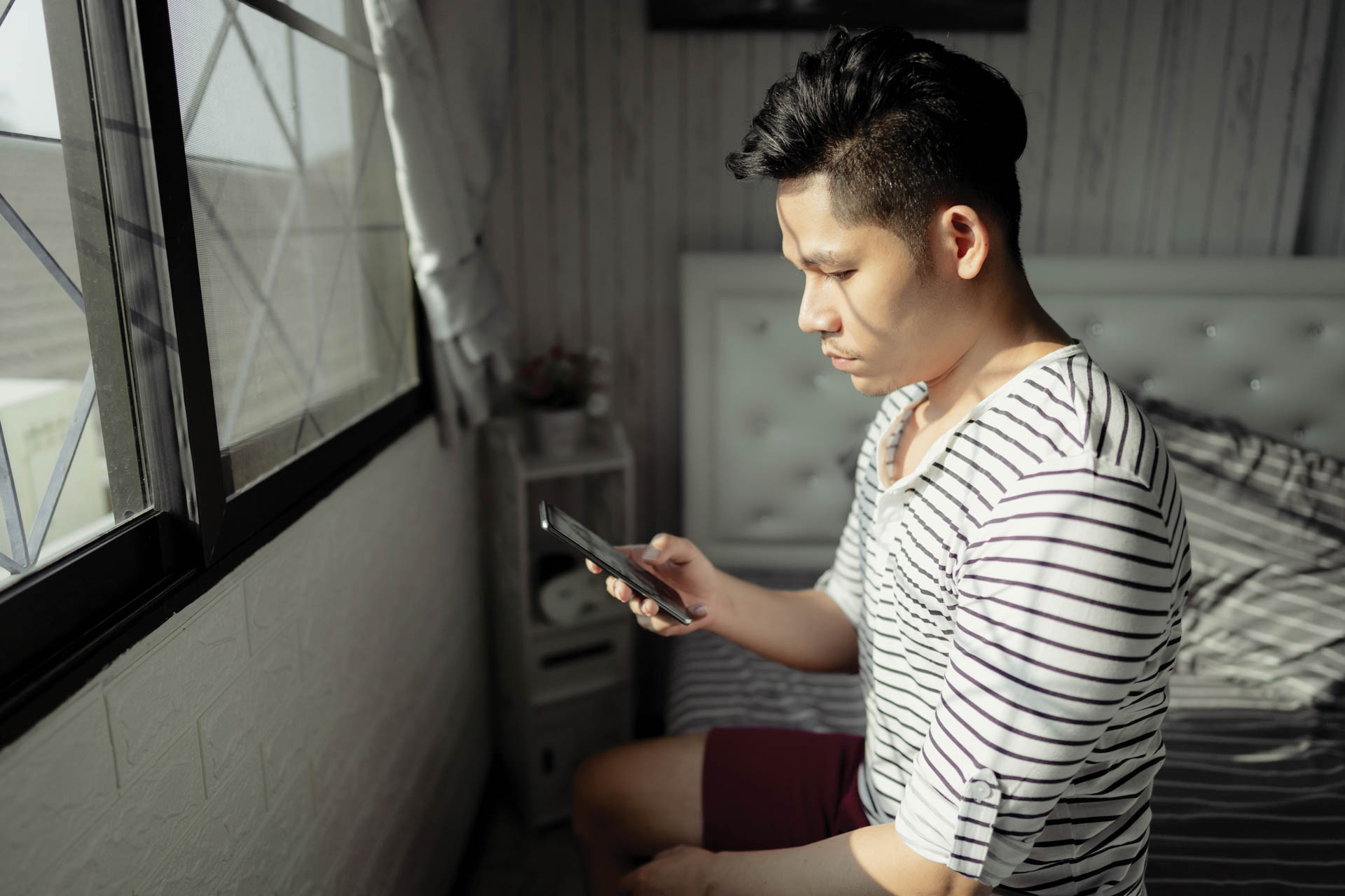 Adult man sitting on his bed, scrolling on his phone.